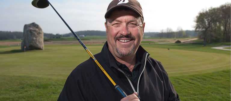 man with black jacket, shirt, and hat is smiling into the camera, with a golf club in his hand