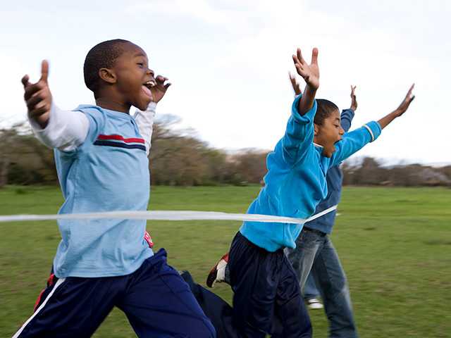 Three African American boys, in blue shirts and long pants, running outside with arms raised in the air