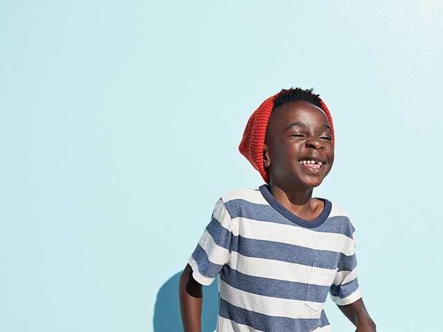 Young African American boy in blue and white striped shirt smiling while standing in front of light blue wall
