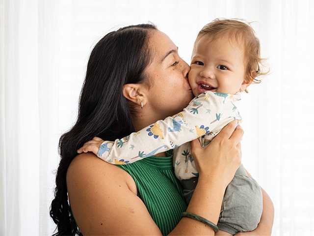 Young woman with shoulder length dark brown hair holding a baby and kissing it on the cheek