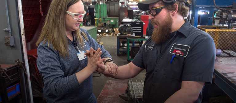 female medical professional examines hand and wrist function in a factory of worker, both wearing protective glasses