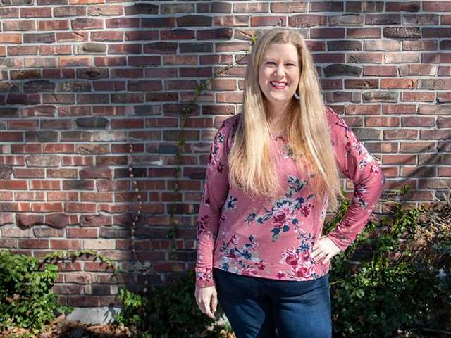 Woman with long blonde hair, wearing a pink shirt, standing in front of a brick wall