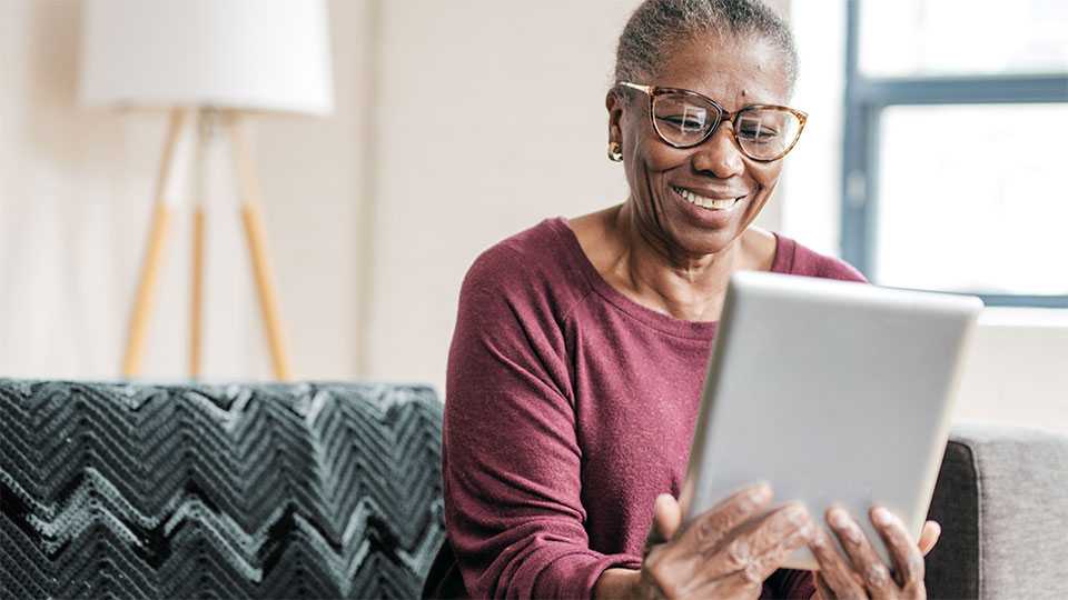 black woman with grey hair sitting, smiling and looking at a tablet in her hands